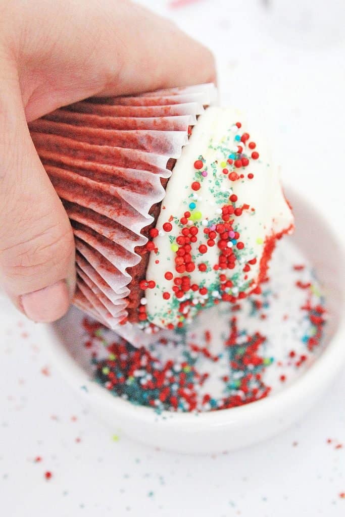 Red cupcake with white icing and multicolored sprinkles being dipped into bowl of sprinkles.