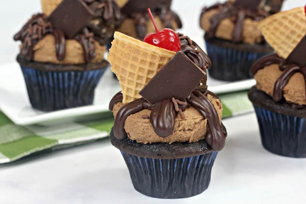 Chocolate cupcakes topped with cookie dough, ganache, hershey bar and waffle cone piece sitting on countertop.
