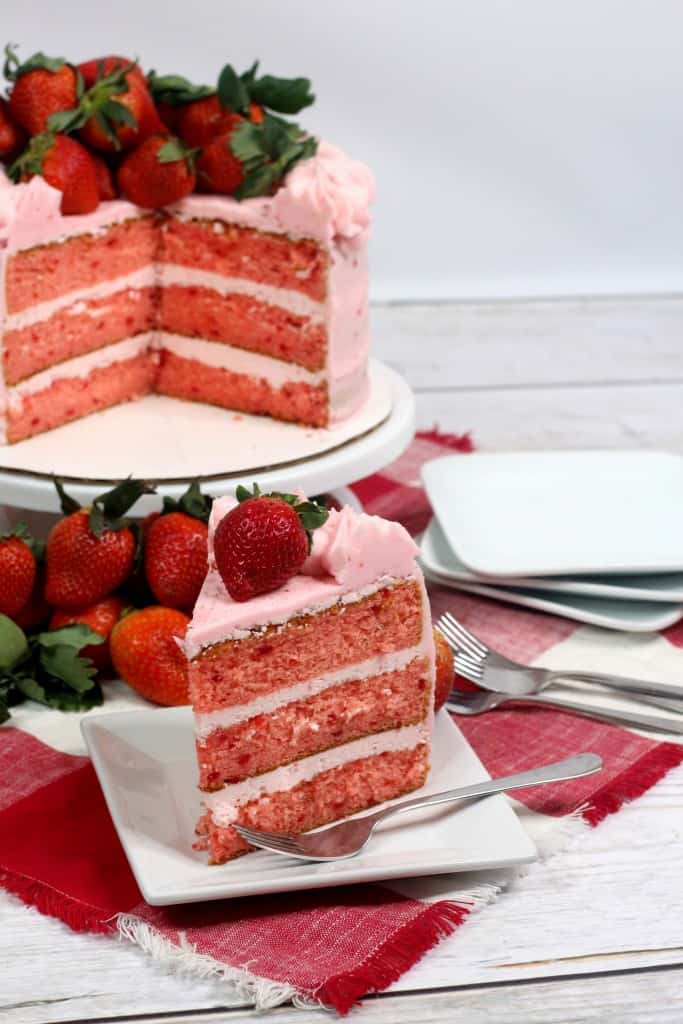 Slice of 3-layer strawberry cake sitting on white plate on wooden countertop with fork on the plate.
